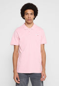 Junger Mann mit lockigem Haar, der ein hellrosa kurzärmeliges Poloshirt und dunkle Hosen trägt, steht vor einem schlichten Hintergrund.
