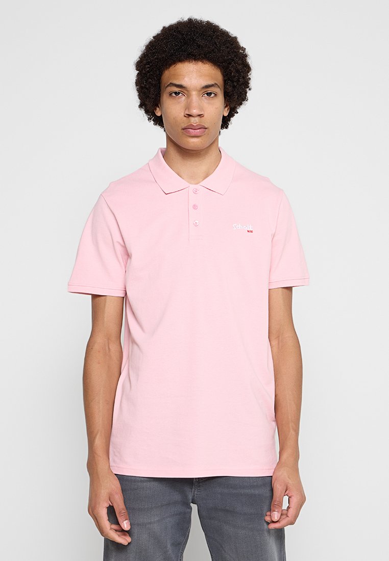 Junger Mann mit lockigem Haar, der ein hellrosa kurzärmeliges Poloshirt und dunkle Hosen trägt, steht vor einem schlichten Hintergrund.