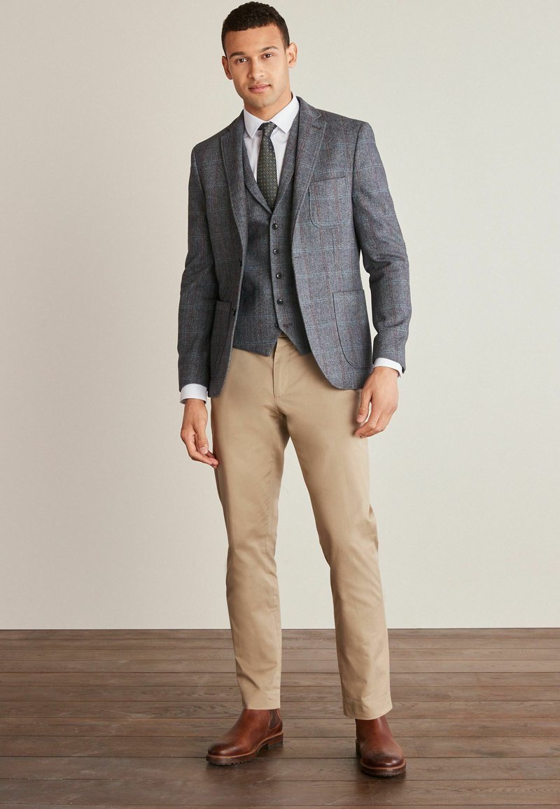 Grau karierter Blazer mit Reverskragen über einer passenden Weste, kombiniert mit beigen Hosen und braunen Lederstiefeln. Glatte Textur und taillierte Passform.