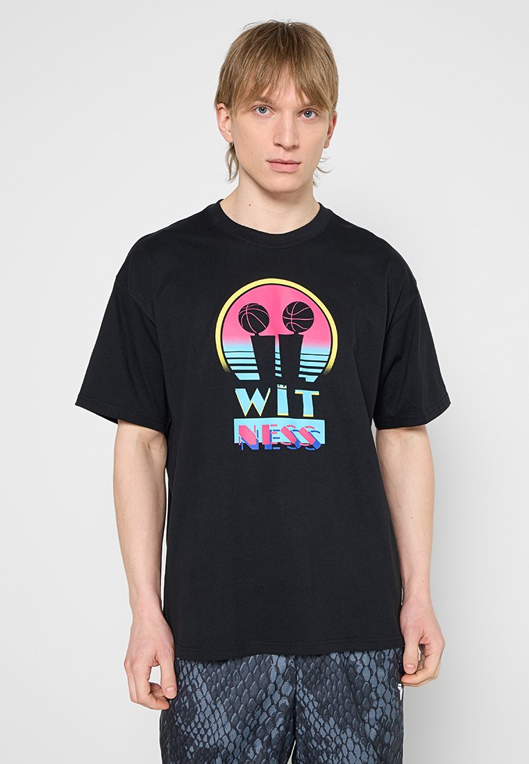 Junger Mann trägt ein schwarzes T-Shirt mit buntem Grafikdruck von zwei Basketballkörben und dem Schriftzug "WITNESS" über gemusterten Shorts.