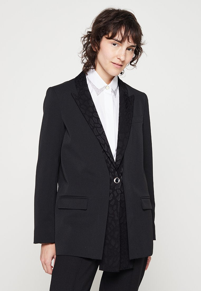 Schwarzer Blazer mit einem gemusterten Satinklappen und einem einzelnen silbernen Knopf. Verfügt über zwei Fronttaschen und eine taillierte Passform. Darunter ein weißes Hemd.