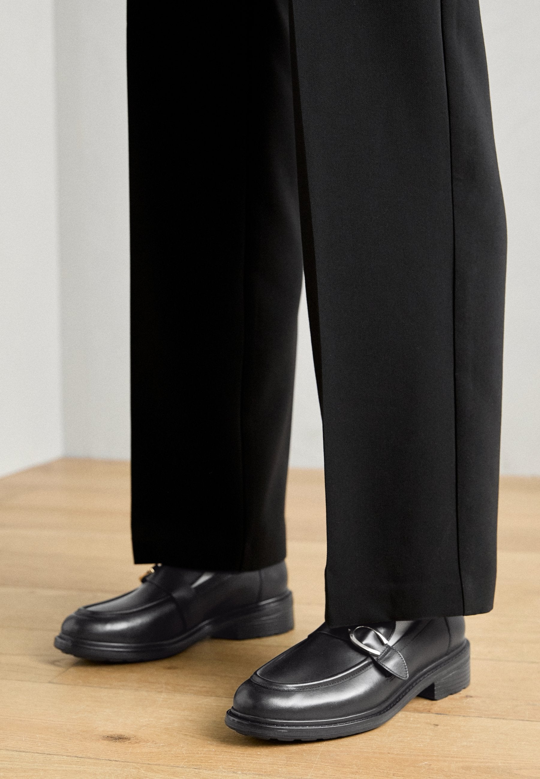 Schwarze Lederloafers mit abgerundeter Spitze, dekorativem Riemen und niedrigem Absatz, kombiniert mit schwarzen Hosen, auf einem Holzboden.