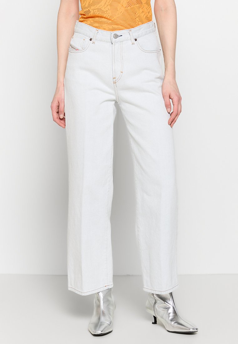 Weiße weit geschnittene Jeans mit hoher Taille und Vordertaschen. Kupferfarbene Nähte und ein Metallknopf vervollständigen das Design. Tragen mit silbernen Knöchelboots.