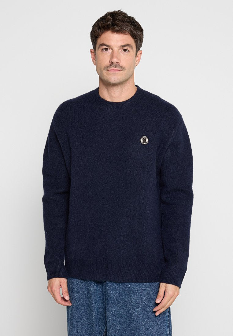 Dunkelblauer Strickpullover mit rundem Halsausschnitt, gerippten Ärmeln und einem kleinen gestickten Logo auf der Brust. Weiche Textur, entspannte Passform.