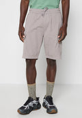 Mann trägt hellgraue Cargoshorts, grünes T-Shirt, graue gemusterte Socken und schwarze und graue Sportschuhe, steht auf einem weißen Boden.