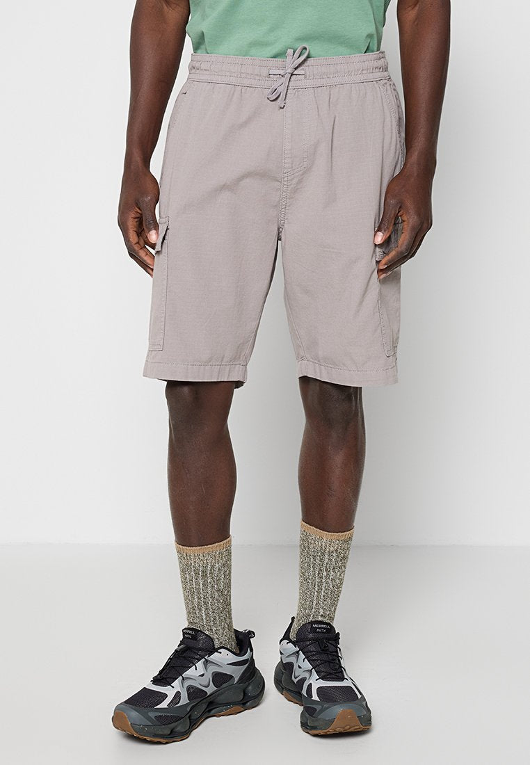 Mann trägt hellgraue Cargoshorts, grünes T-Shirt, graue gemusterte Socken und schwarze und graue Sportschuhe, steht auf einem weißen Boden.