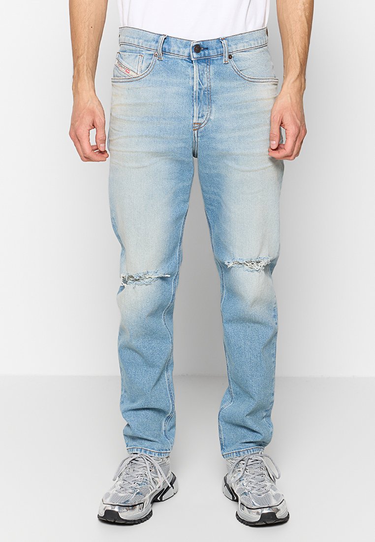 Hellblaue Denim-Jeans mit lockerer Passform, die an den Knien aufgerissene Stellen und dezente Verwaschungen aufweisen. Kombiniert mit grauen Sportschuhen.