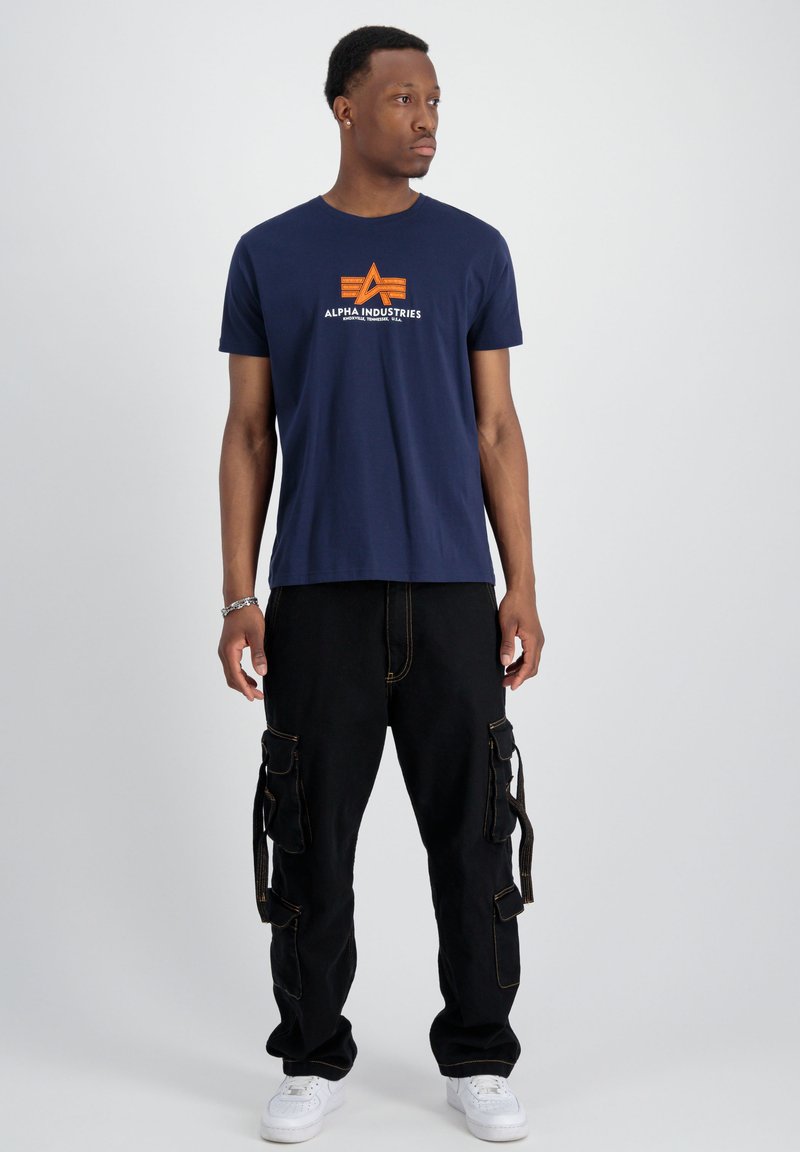 Ein marineblaues T-Shirt mit einem zentralen orange-weißen Logo. Kombiniert mit schwarzen Cargo-Hosen mit mehreren Taschen und weißen Sneakern.