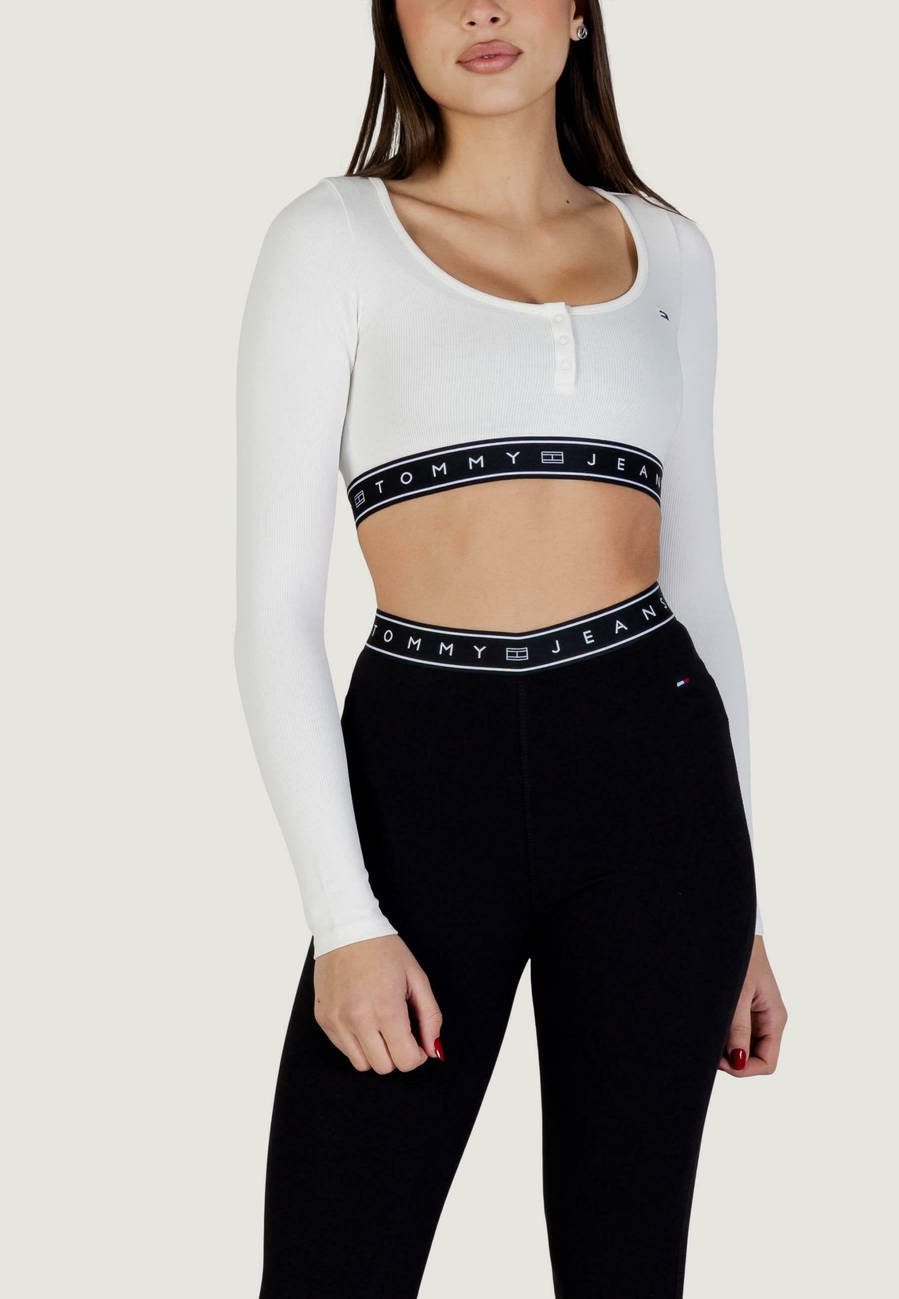 Weißes, geripptes Crop-Top mit langen Ärmeln und Knopfdetails sowie einem schwarzen elastischen Bund mit dem "TOMMY JEANS"-Logo; kombiniert mit hoch taillierten schwarzen Leggings.