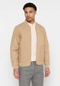 Beige gesteppte Bomberjacke mit einem runden Kragen, Frontreißverschluss und aufgesetzten Taschen. Getragen über einem hellen Polohemd und grauen Hosen.