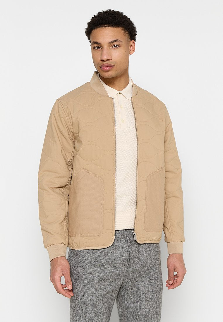 Beige gesteppte Bomberjacke mit einem runden Kragen, Frontreißverschluss und aufgesetzten Taschen. Getragen über einem hellen Polohemd und grauen Hosen.