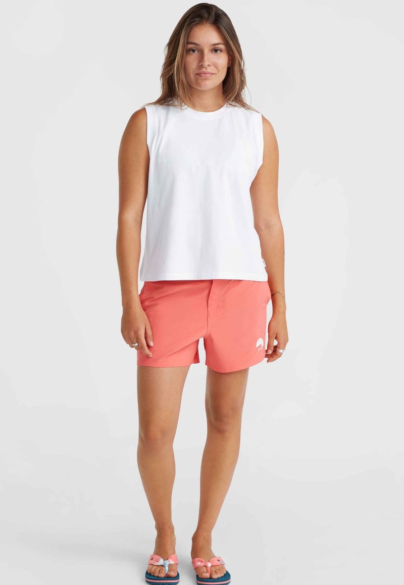 Weißes ärmelloses Top kombiniert mit korallenfarbenen Shorts mit Logo. Das Model trägt pinke Sandalen mit Schleifen-Akzenten. Einfacher, lässiger Strandlook.