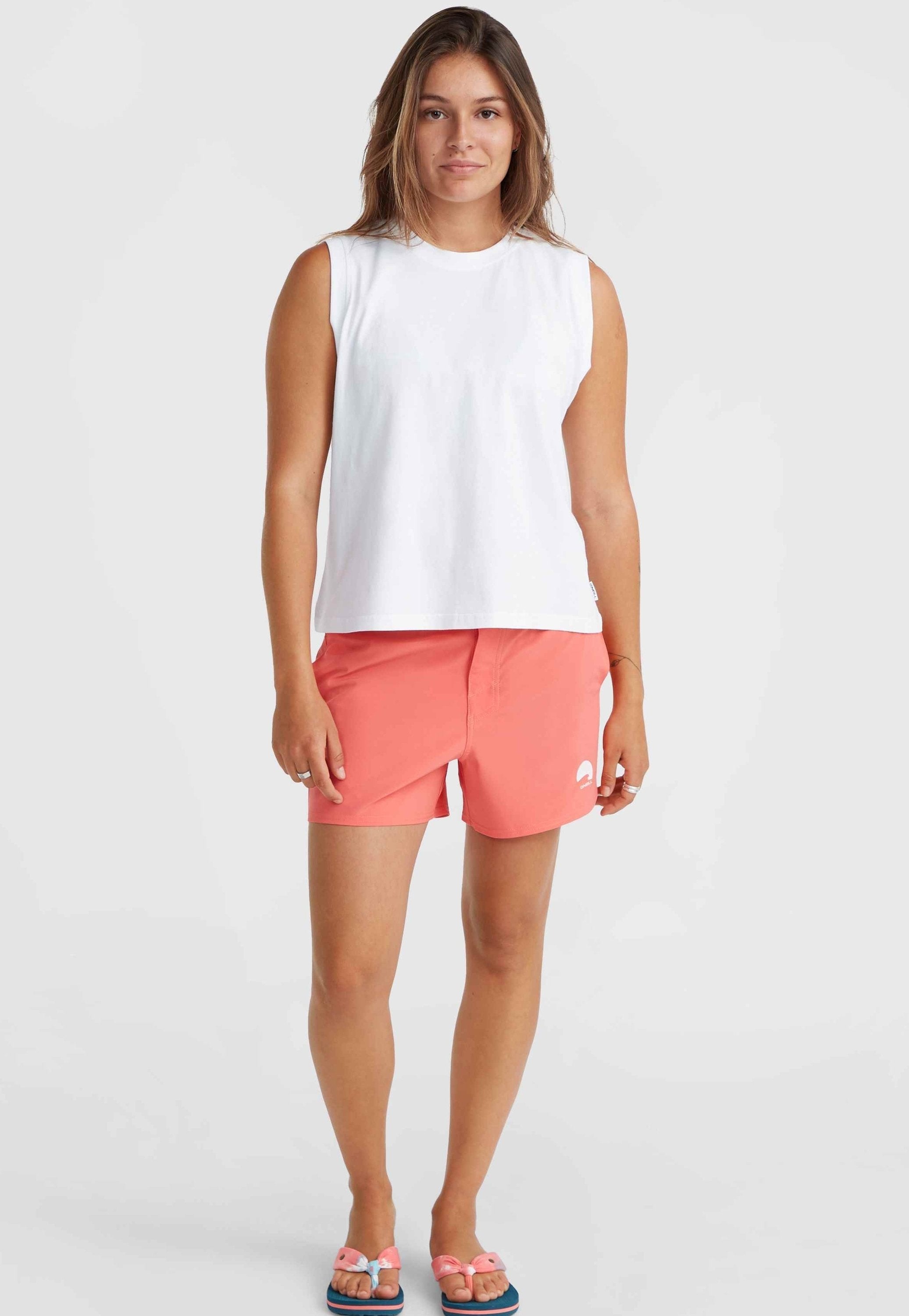 Weißes ärmelloses Top kombiniert mit korallenfarbenen Shorts mit Logo. Das Model trägt pinke Sandalen mit Schleifen-Akzenten. Einfacher, lässiger Strandlook.