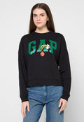 Junge Frau mit langen braunen Haaren, die ein schwarzes GAP-Sweatshirt mit grünen Buchstaben und einem Cartoon-Detail trägt, kombiniert mit blauen Jeans.
