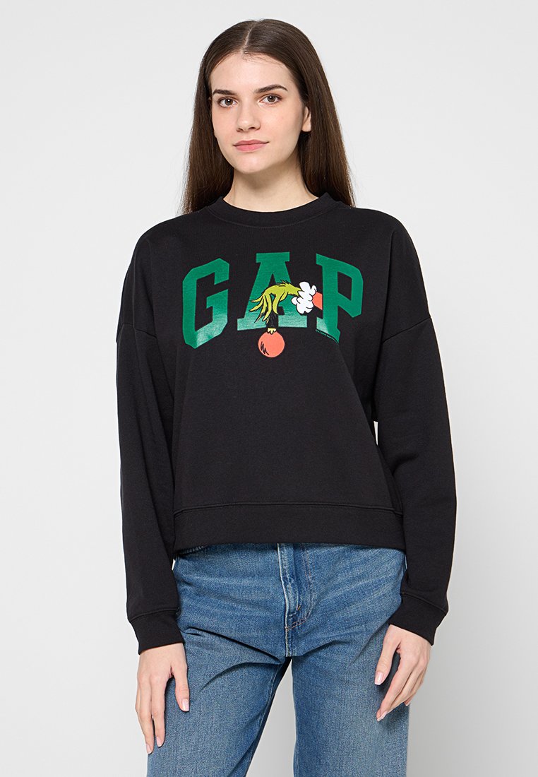 Junge Frau mit langen braunen Haaren, die ein schwarzes GAP-Sweatshirt mit grünen Buchstaben und einem Cartoon-Detail trägt, kombiniert mit blauen Jeans.