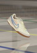 Weißer und blauer Nike Tiempo Hallenfußballschuh schwebt über einem Hallenboden mit mehrfarbigen Spielfeldlinien.