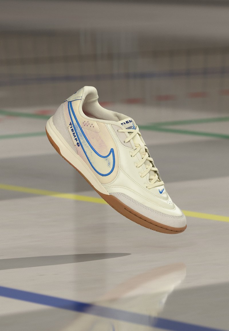 Weißer und blauer Nike Tiempo Hallenfußballschuh schwebt über einem Hallenboden mit mehrfarbigen Spielfeldlinien.