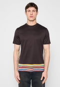 Mann trägt ein schwarzes, kurzärmliges T-Shirt mit mehrfarbigen horizontalen Streifen am Saum, kombiniert mit dunklen Jeans, steht vor einer weißen Wand.