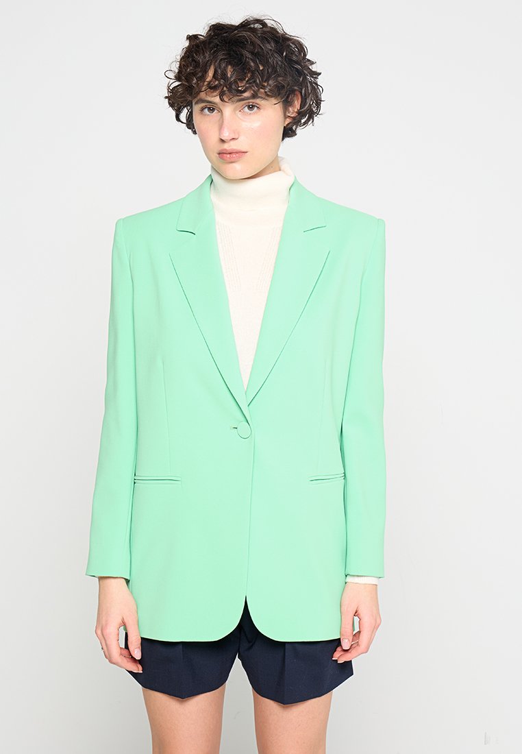 Minzgrüner, taillierter Blazer mit einem einzigen Knopfverschluss, zwei vorderen Taschen und glatter Textur, getragen über einem hellen Rollkragenpullover und Shorts.