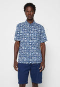 Mann mit blauem Kurzarmhemd mit weißen geometrischen und Blättermustern und marineblauen knielangen Shorts.