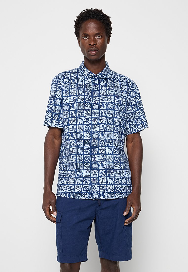 Mann mit blauem Kurzarmhemd mit weißen geometrischen und Blättermustern und marineblauen knielangen Shorts.