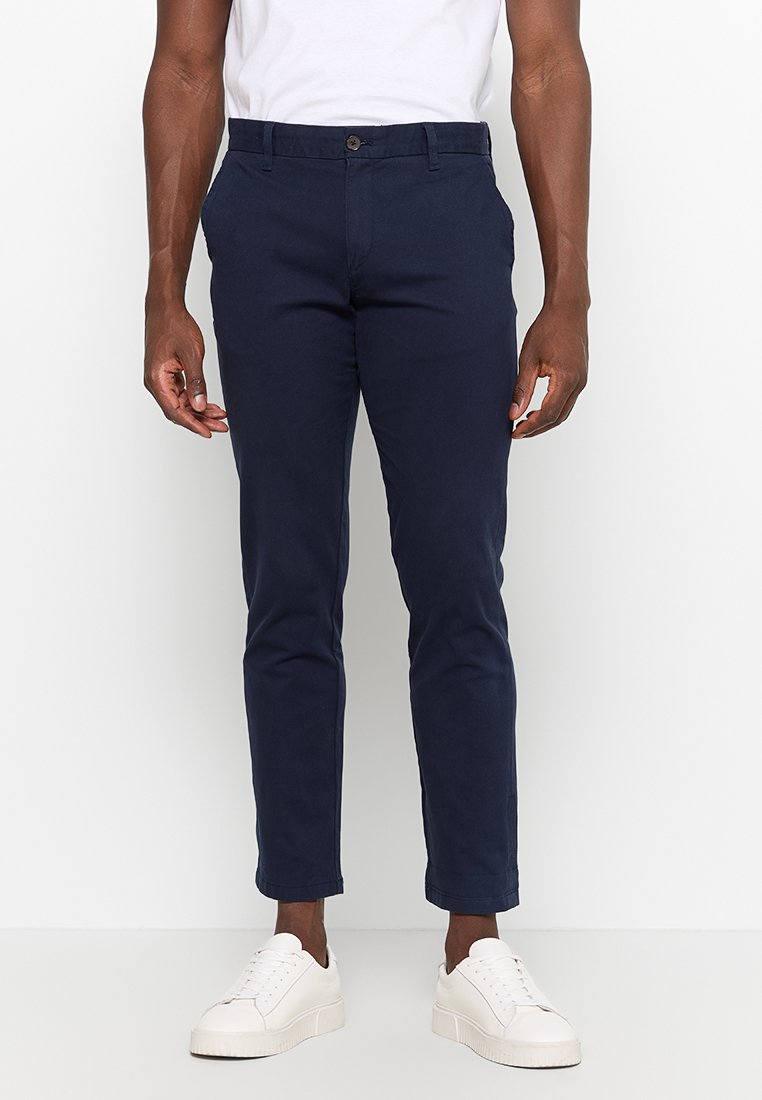 Marineblaue Hosen aus glattem Baumwollstoff, Slim Fit, mit Gürtelbund und Reißverschluss vorne, kombiniert mit weißen Sneakers.