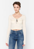 Cremigere, gerippte Langarm-Crop-Top mit V-Ausschnitt, zwei Fronttaschen und Reißverschluss. Kombiniert mit hoch geschnittenen blauen Jeans.