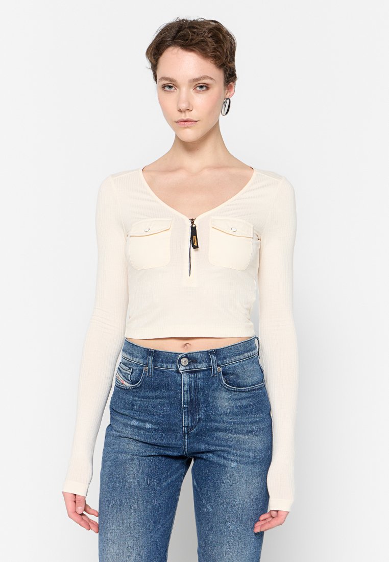 Cremigere, gerippte Langarm-Crop-Top mit V-Ausschnitt, zwei Fronttaschen und Reißverschluss. Kombiniert mit hoch geschnittenen blauen Jeans.