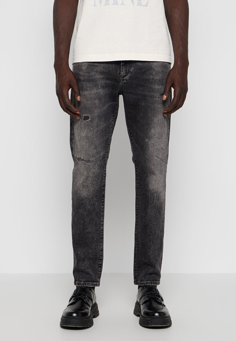 Schwarze Jeans aus Denim mit einem ausgebleichten und lädierten Textur, mit Rissen an den Knien und einem schmalen Schnitt. Getragen mit schwarzen Schnürschuhen.