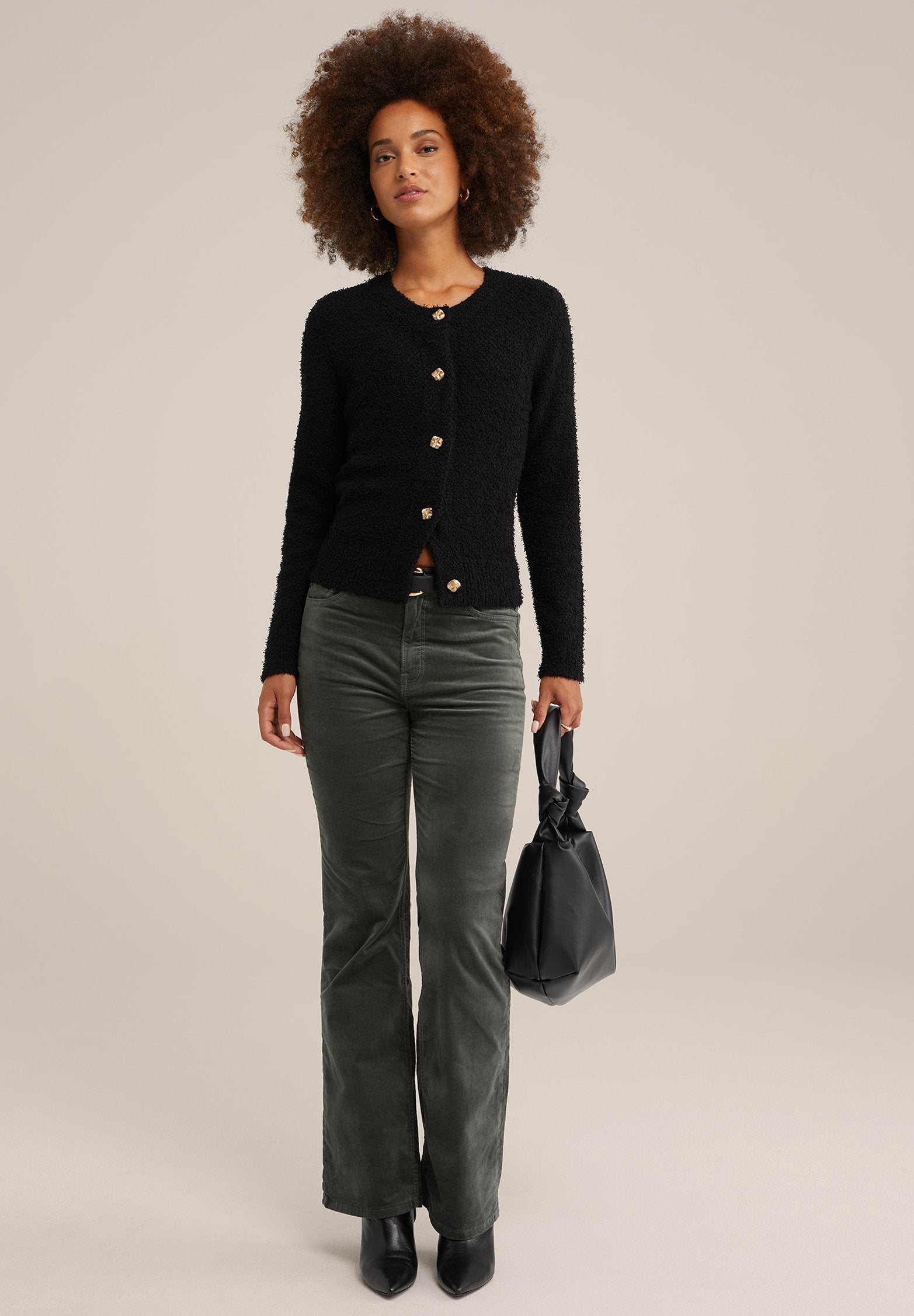 Schwarze, flauschige Strickjacke mit goldenen Knöpfen, kombiniert mit dunkelgrünen, ausgestellten Cordhosen und einer schwarzen Handtasche. Schwarze Stiefeletten mit Absatz runden das Outfit ab.