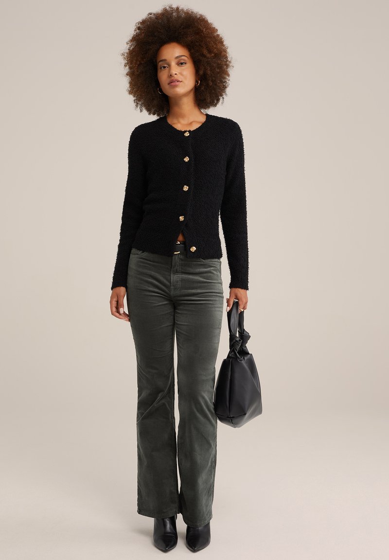 Schwarze, flauschige Strickjacke mit goldenen Knöpfen, kombiniert mit dunkelgrünen, ausgestellten Cordhosen und einer schwarzen Handtasche. Schwarze Stiefeletten mit Absatz runden das Outfit ab.