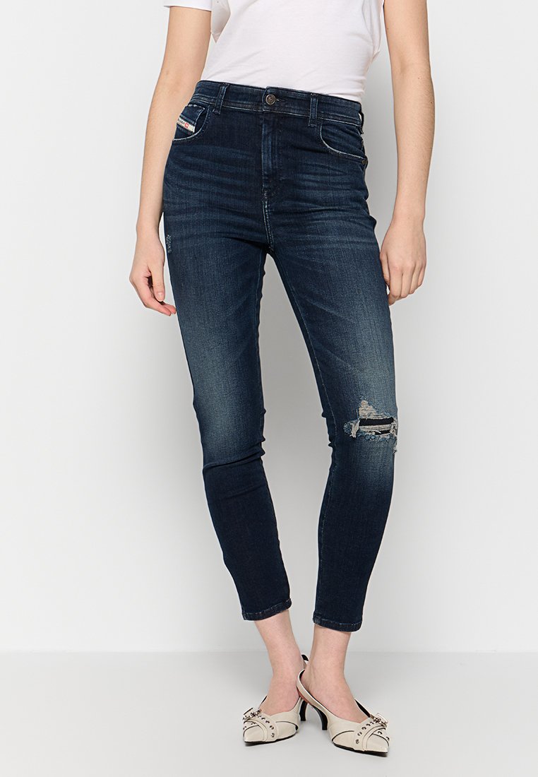 Schlanke, dunkelblaue Denim-Jeans mit hoher Taille, ausgestattet mit einem beschädigten Knie mit einem Riss, ergänzt durch weiße Nähte und einen Knopfverschluss.
