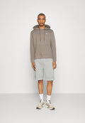 Grauer Hoodie mit einer Fronttasche und Kordelzug, kombiniert mit hellgrauen Shorts. Die Schuhe zeigen eine Mischung aus Beige- und Cremetönen.