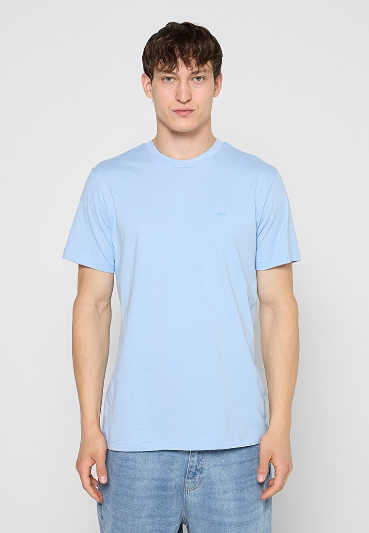 Junger Mann, der ein hellblaues T-Shirt mit kurzen Ärmeln und blaue Jeans trägt und vor einem einfarbigen grauen Hintergrund steht.