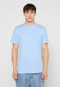 Junger Mann, der ein hellblaues T-Shirt mit kurzen Ärmeln und blaue Jeans trägt und vor einem einfarbigen grauen Hintergrund steht.