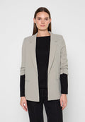 Frau mit glatten braunen Haaren trägt einen hellgrauen Blazer über einem schwarzen Langarmtop und schwarze Hose, steht vor schlichtem hellem Hintergrund.