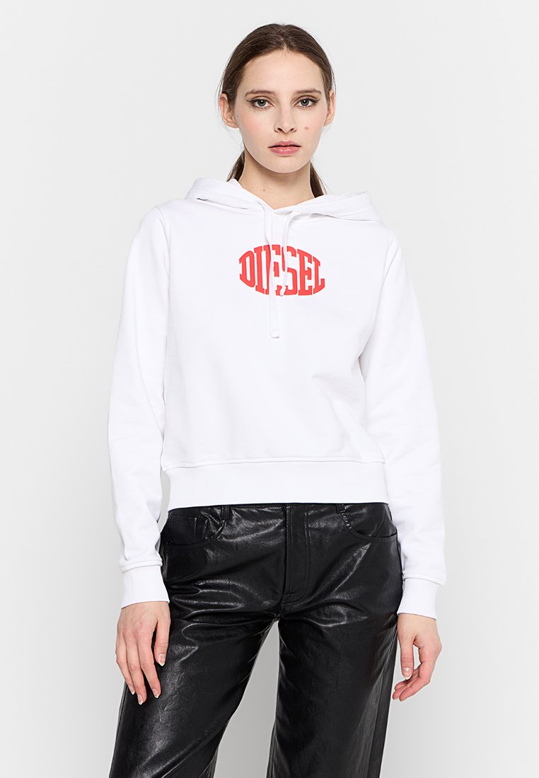 Frau, die einen weißen Hoodie mit rotem "DIESEL"-Logo und schwarze Lederhosen trägt, und vor einem schlichten weißen Hintergrund steht.