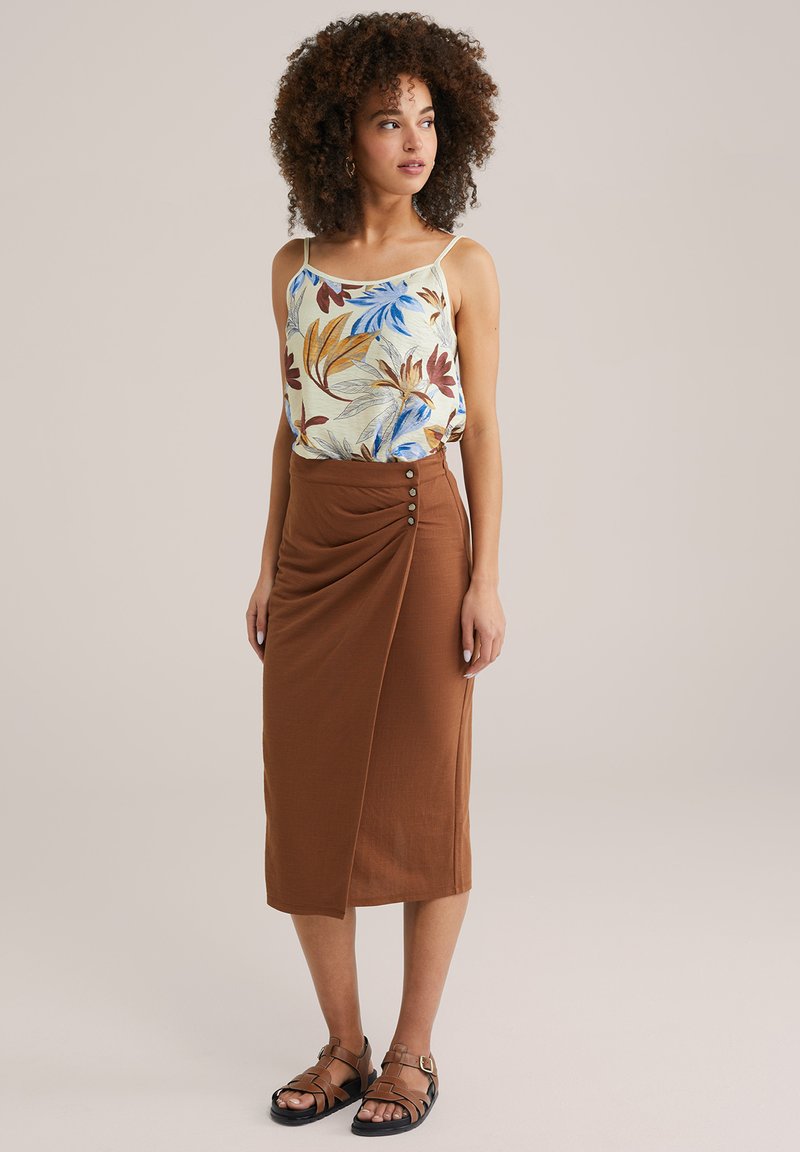 Leichtes Tanktop mit floralen Mustern in Creme, kombiniert mit einem braunen, knielangen, drapierten Rock mit Knopfdetails. Schwarze Sandalen mit Riemchen.