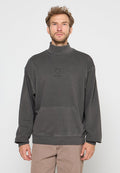 Mann mit lockigem Haar, der einen dunkelgrauen Rollkragenpullover mit Fronttasche und beigen Hosen trägt, steht vor einem schlichten weißen Hintergrund.