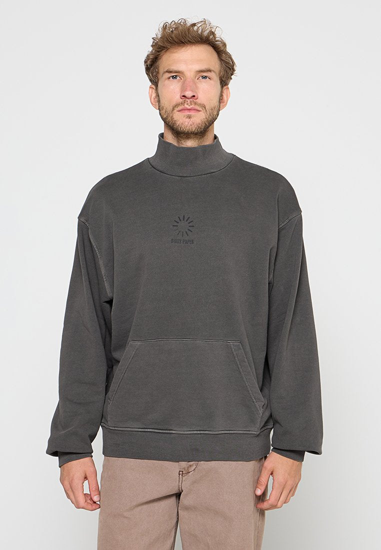 Mann mit lockigem Haar, der einen dunkelgrauen Rollkragenpullover mit Fronttasche und beigen Hosen trägt, steht vor einem schlichten weißen Hintergrund.