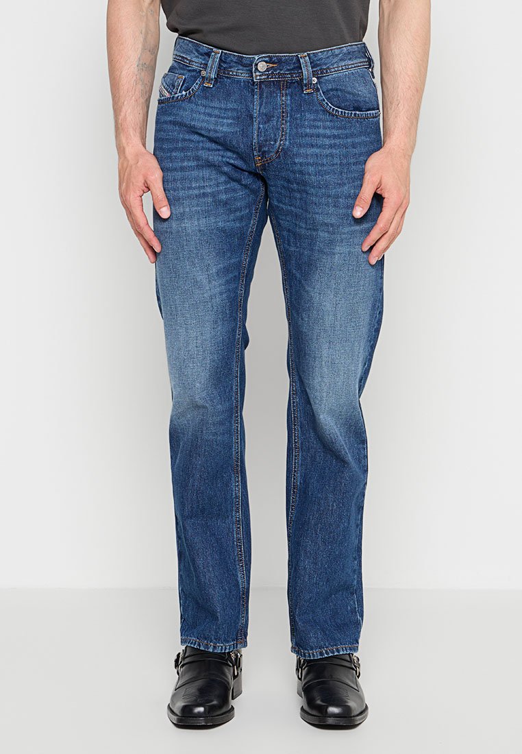 Mann trägt blaue Jeans mit geradem Schnitt und schwarze Lederschuhe, steht vor einem schlichten weißen Hintergrund.