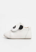 Weißer Babyschuh mit schwarzem, augenförmigem Design auf der verstellbaren Klettlasche, weicher Sohle und gesteppten Details.