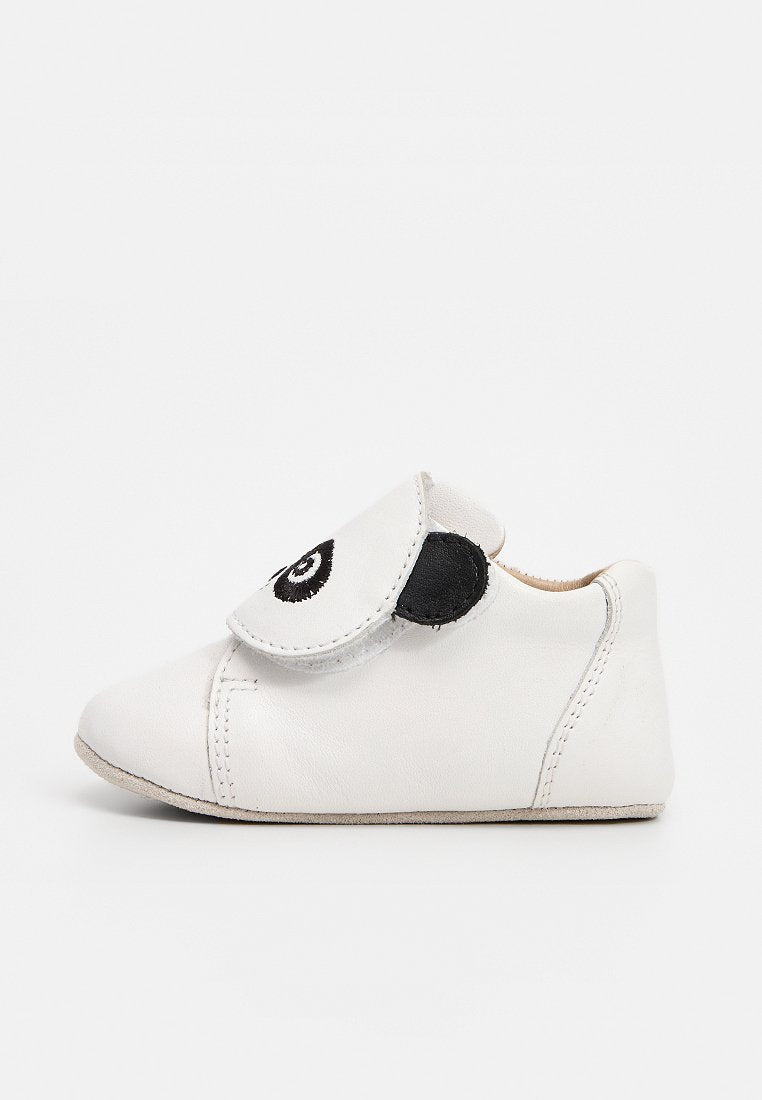 Weißer Babyschuh mit schwarzem, augenförmigem Design auf der verstellbaren Klettlasche, weicher Sohle und gesteppten Details.
