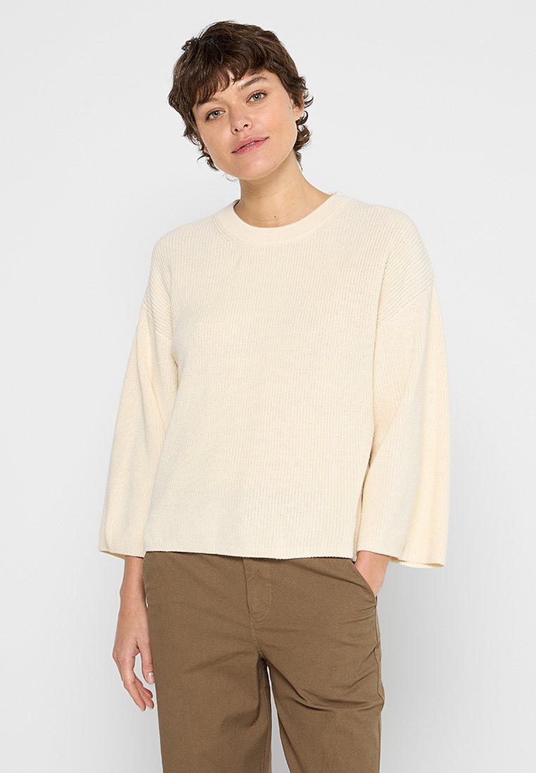 Frau mit kurzen braunen Haaren, die einen cremefarbenen, langärmeligen Strickpullover und braune Hosen trägt und mit einer Hand in der Tasche vor einem weißen Hintergrund steht.