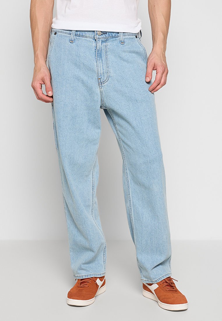 Hellblaue Denim-Jeans mit lockerer Passform, ausgestattet mit einem Reißverschluss und fünf Taschen. Sie werden mit braunen Sneakers getragen, der Stoff ist glatt und klassisch.