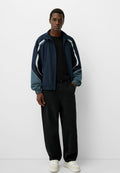 Navy-blaues Windbreaker mit weißen und türkisfarbenen Akzenten, ausgestattet mit elastischen Bündchen und einer lässigen Passform, kombiniert mit schwarzen Hosen und weißen Turnschuhen.