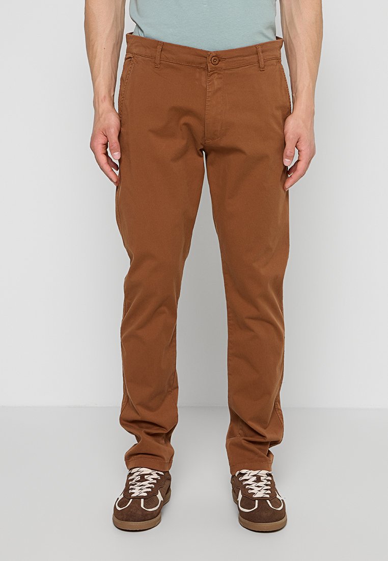 Mann trägt braune Freizeithose und braune Sneakers mit weißen Schnürsenkeln, steht vor einem schlichten hellen Hintergrund.