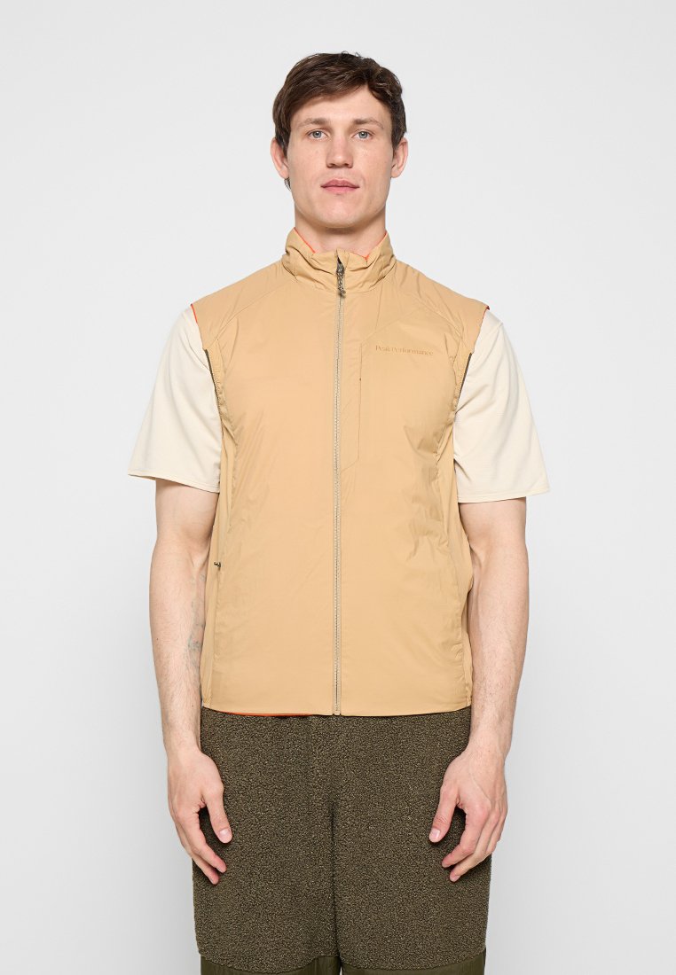 Mann trägt beige ärmellose Weste mit Reißverschluss über weißem kurzärmligem Hemd und strukturierten braunen Shorts, steht vor schlichtem hellem Hintergrund.