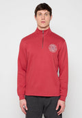 Mann trägt einen roten Pullover mit Viertel-Reißverschluss und einem weißen, runden Logo "Worldwide Established 1989" auf der linken Brust, kombiniert mit dunkler Hose.
