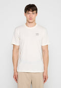 Junger Mann mit kurzen braunen Haaren, der ein schlichtes weißes T-Shirt mit dem Schriftzug „MCXX“ auf der Brust und beigefarbene Hosen vor einem weißen Hintergrund trägt.